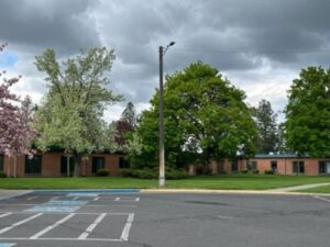 An asphalt parking lot with handicap parking spaces that leads to a one story brick building with many windows and surrounded by tall deciduous trees and shrubs.