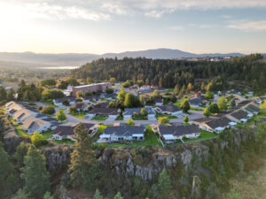 An aerial view of the Olympus of Spokane Valley of Olympus Living campus. Steep rocks and pine trees lead into well-kept yards of green grass, flower beds and three streets of twinhomes. A four-story brick building sits at the far end of the campus. Behind the building is a large grove of pine trees, more homes and a mountain range.